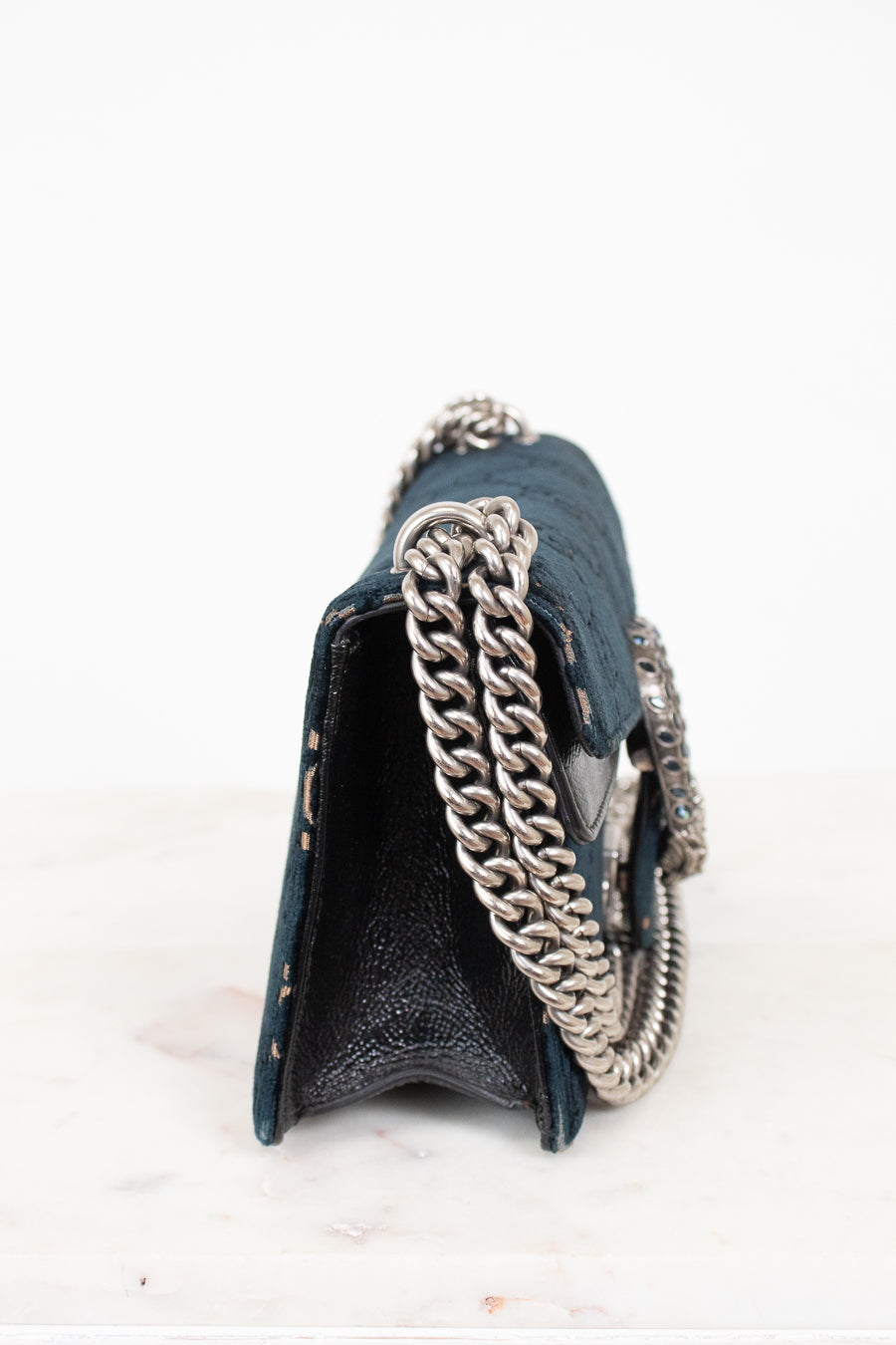 A close-up side view of a dark teal textured leather handbag with a chunky silver chain strap, resting on a white marble surface against a white background.
