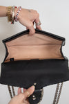 A close-up of a black leather handbag with a tan interior lining, held open by two hands wearing gold jewelry against a plain light background.