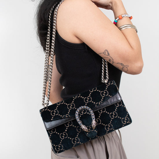 A woman wearing a black tank top and beige pants carries a teal Gucci shoulder bag with the brand's signature GG monogram pattern and a silver-tone dionysus clasp, shown from a side-back angle against a plain light gray background.