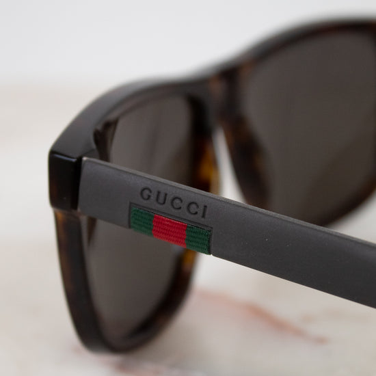 Close-up of Gucci sunglasses with dark brown tortoiseshell frames, dark lenses, and the signature green and red web stripe detail on the temple arm.