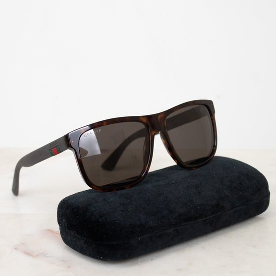 A pair of square-frame sunglasses with dark brown tortoiseshell frames and dark lenses, resting on a black velvet case against a white background.