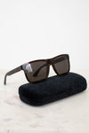 A pair of square-frame sunglasses with dark brown tortoiseshell frames and dark lenses, resting on a black velvet case against a white background.