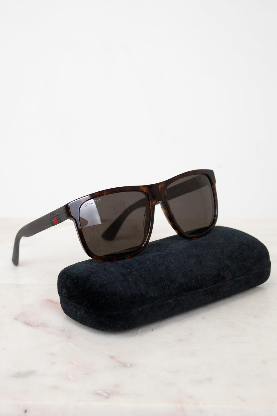 A pair of square-frame sunglasses with dark brown tortoiseshell frames and dark lenses, resting on a black velvet case against a white background.