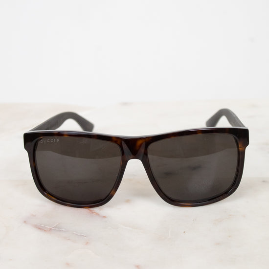 A pair of square-frame sunglasses with dark brown tortoiseshell frames and dark gray lenses, resting on a white marble surface against a white background.
