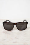 A pair of square-frame sunglasses with dark brown tortoiseshell frames and dark gray lenses, resting on a white marble surface against a white background.