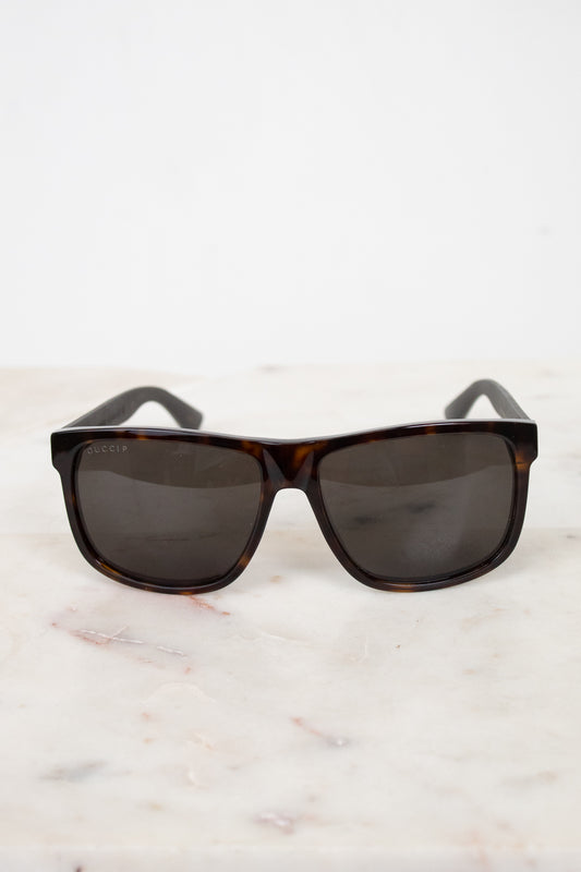 A pair of square-frame sunglasses with dark brown tortoiseshell frames and dark gray lenses, resting on a white marble surface against a white background.