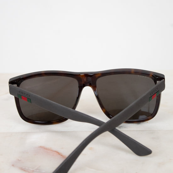 Gucci sunglasses with a dark brown and black tortoiseshell frame, dark lenses, and matte black temples featuring the signature red and green web stripe logo, resting on a light marble surface against a white background.