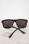 Gucci sunglasses with a dark brown and black tortoiseshell frame, dark lenses, and matte black temples featuring the signature red and green web stripe logo, resting on a light marble surface against a white background.