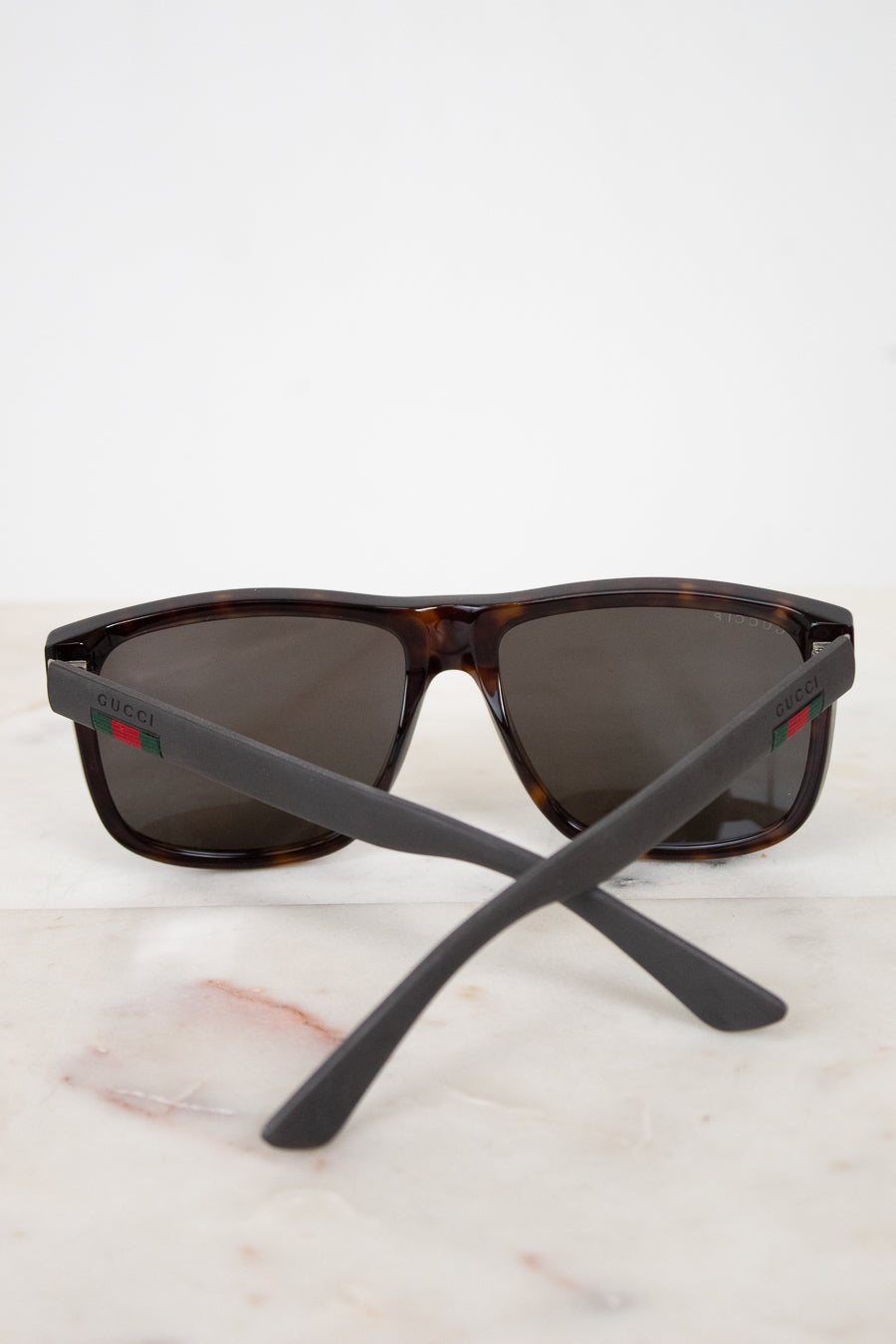Gucci sunglasses with a dark brown and black tortoiseshell frame, dark lenses, and matte black temples featuring the signature red and green web stripe logo, resting on a light marble surface against a white background.