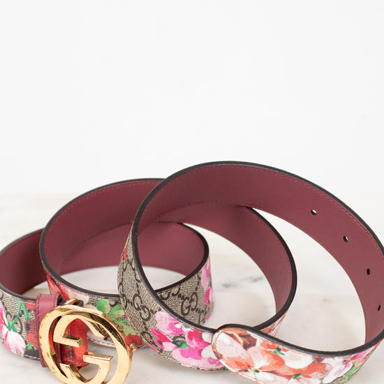 A rolled-up designer belt on a light marble surface against a white background. Belt is featuring the classic brown GG Supreme monogram with a gold-tone interlocking G buckle, and the other features a vibrant pink, red, and white floral print.