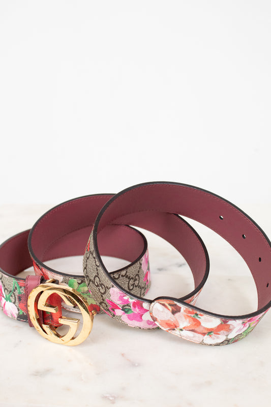 A rolled-up designer belt on a light marble surface against a white background. Belt is featuring the classic brown GG Supreme monogram with a gold-tone interlocking G buckle, and the other features a vibrant pink, red, and white floral print.