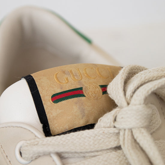 Close-up of a beige Gucci sneaker with cream laces, a tan suede heel tab embossed with 'GUCCI', and the signature green and red  stripe.