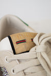 Close-up of a beige Gucci sneaker with cream laces, a tan suede heel tab embossed with 'GUCCI', and the signature green and red  stripe.