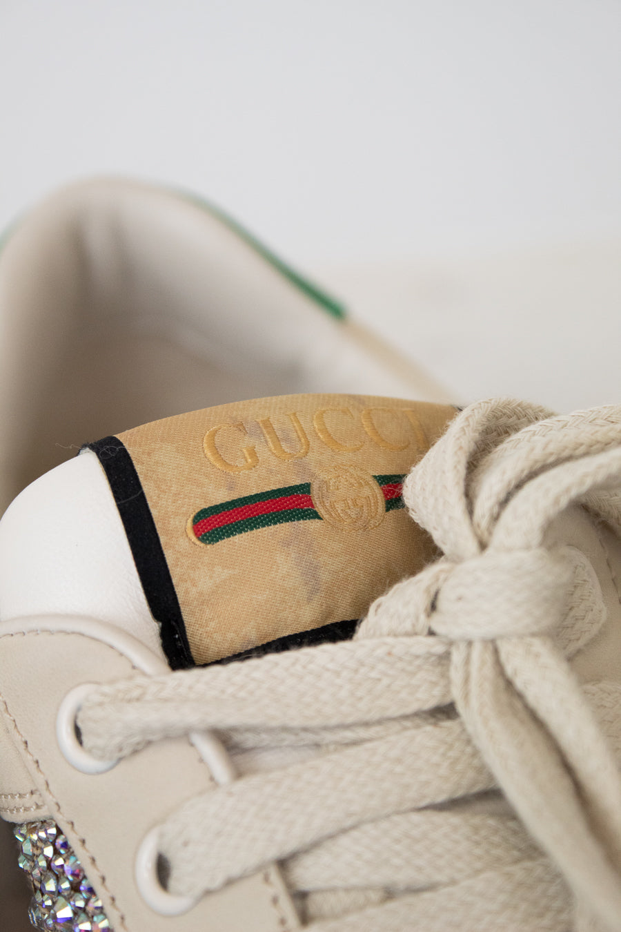 Close-up of a beige Gucci sneaker with cream laces, a tan suede heel tab embossed with 'GUCCI', and the signature green and red  stripe.