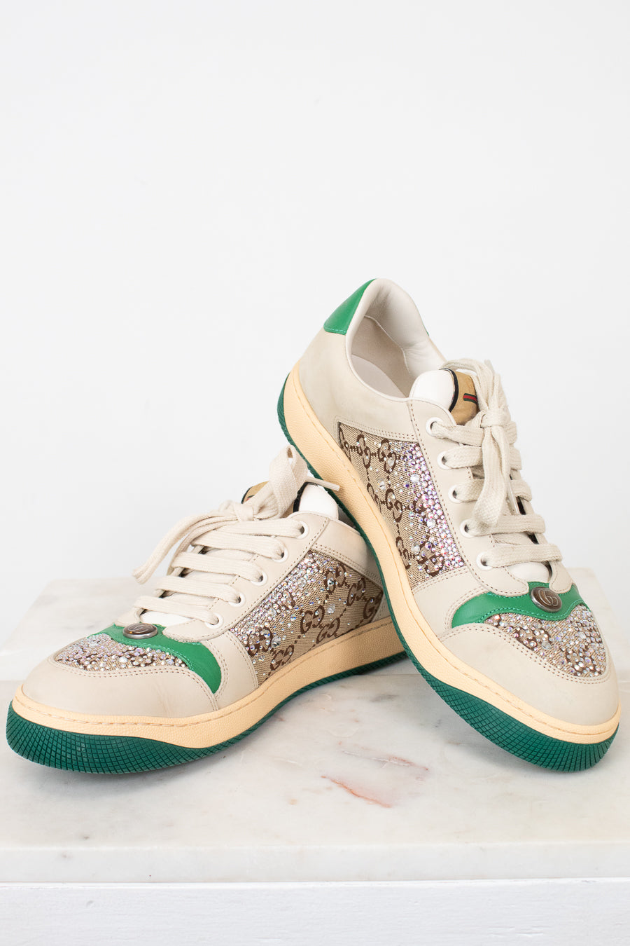A pair of low-top sneakers with beige panels, green accents on the heel and midsole, and a green outsole, displayed on a light marble surface against a white background.
