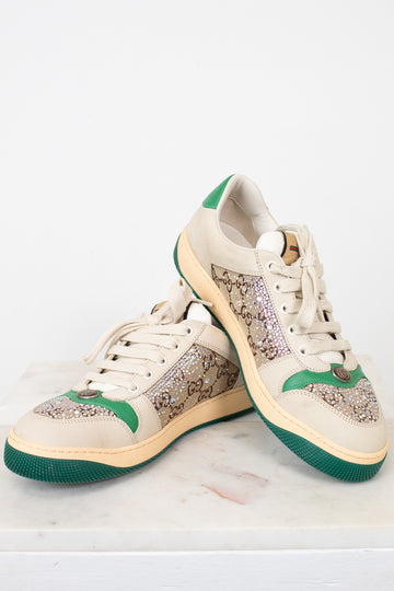 A pair of low-top sneakers with beige panels, green accents on the heel and midsole, and a green outsole, displayed on a light marble surface against a white background.