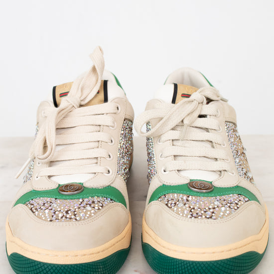 A pair of low-top sneakers in cream canvas with green rubber toe caps and soles, decorated with silver rhinestone embellishments across the eyestay and toe area, resting on a light marble surface against a white background.