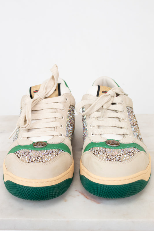 A pair of low-top sneakers in cream canvas with green rubber toe caps and soles, decorated with silver rhinestone embellishments across the eyestay and toe area, resting on a light marble surface against a white background.