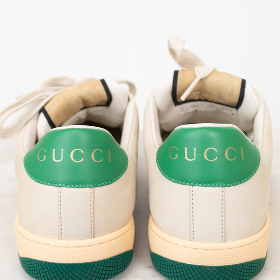 A pair of Gucci low-top sneakers shown from the back, featuring an off-white leather upper, a vibrant green heel tab with gold 'GUCCI' lettering, and a matching green textured outsole.