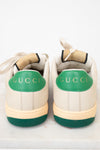 A pair of Gucci low-top sneakers shown from the back, featuring an off-white leather upper, a vibrant green heel tab with gold 'GUCCI' lettering, and a matching green textured outsole.