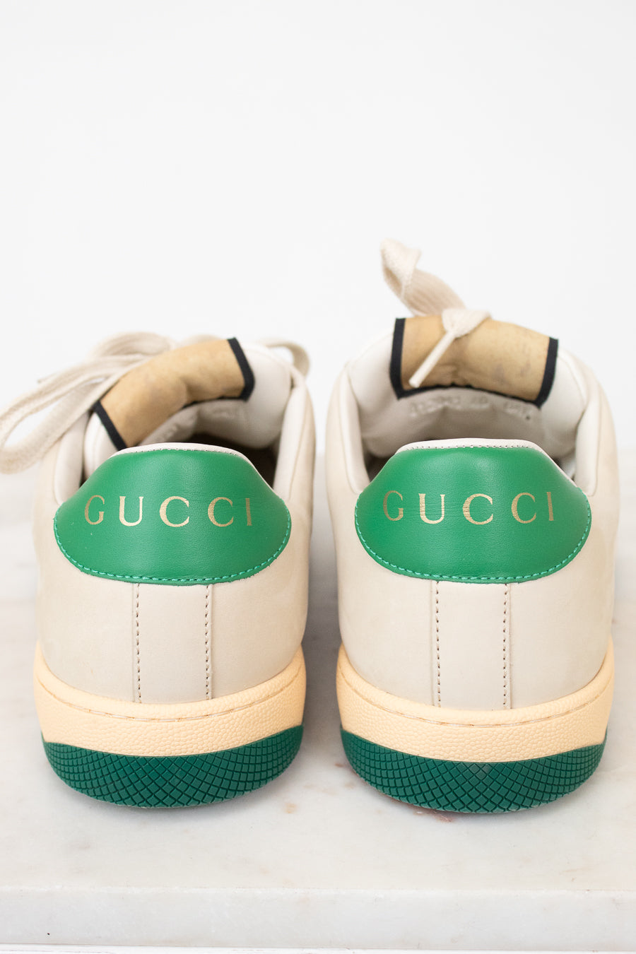 A pair of Gucci low-top sneakers shown from the back, featuring an off-white leather upper, a vibrant green heel tab with gold 'GUCCI' lettering, and a matching green textured outsole.
