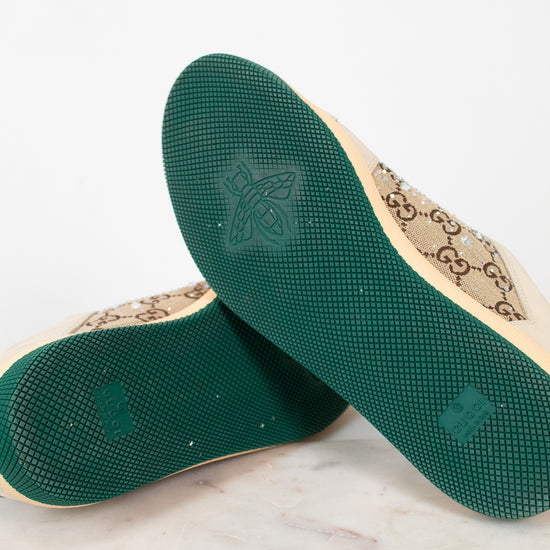 A pair of Gucci low-top canvas shoes shown from the sole side, resting on a white marble surface against a white background. The soles are dark green with a bee logo, and the beige canvas upper features the classic interlocking GG monogram.