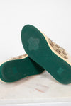 A pair of Gucci low-top canvas shoes shown from the sole side, resting on a white marble surface against a white background. The soles are dark green with a bee logo, and the beige canvas upper features the classic interlocking GG monogram.