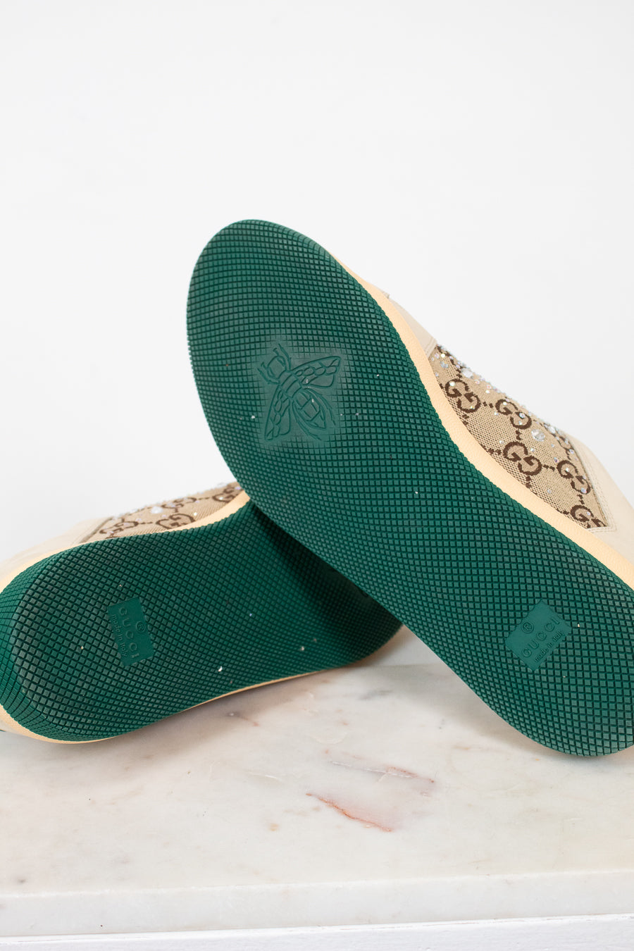A pair of Gucci low-top canvas shoes shown from the sole side, resting on a white marble surface against a white background. The soles are dark green with a bee logo, and the beige canvas upper features the classic interlocking GG monogram.