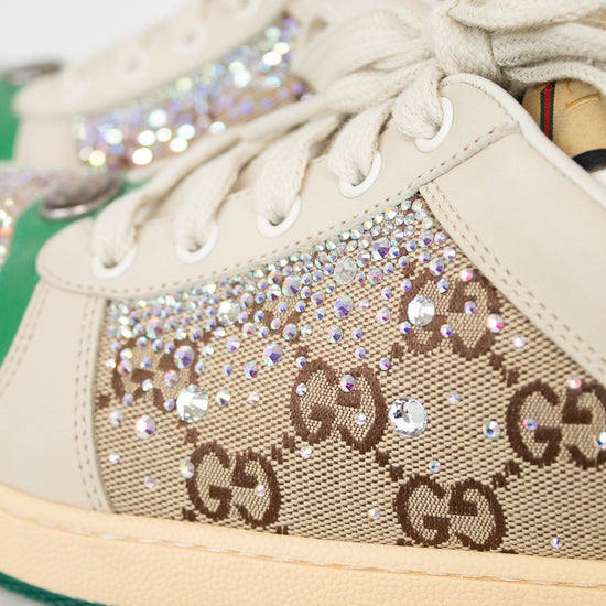 Close-up of a Gucci low-top sneaker featuring beige GG Supreme canvas with brown GG monogram, embellished with iridescent rhinestones and clear crystals, cream laces, a beige rubber sole, and a green stripe along the edge.