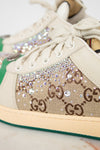 Close-up of a Gucci low-top sneaker featuring beige GG Supreme canvas with brown GG monogram, embellished with iridescent rhinestones and clear crystals, cream laces, a beige rubber sole, and a green stripe along the edge.