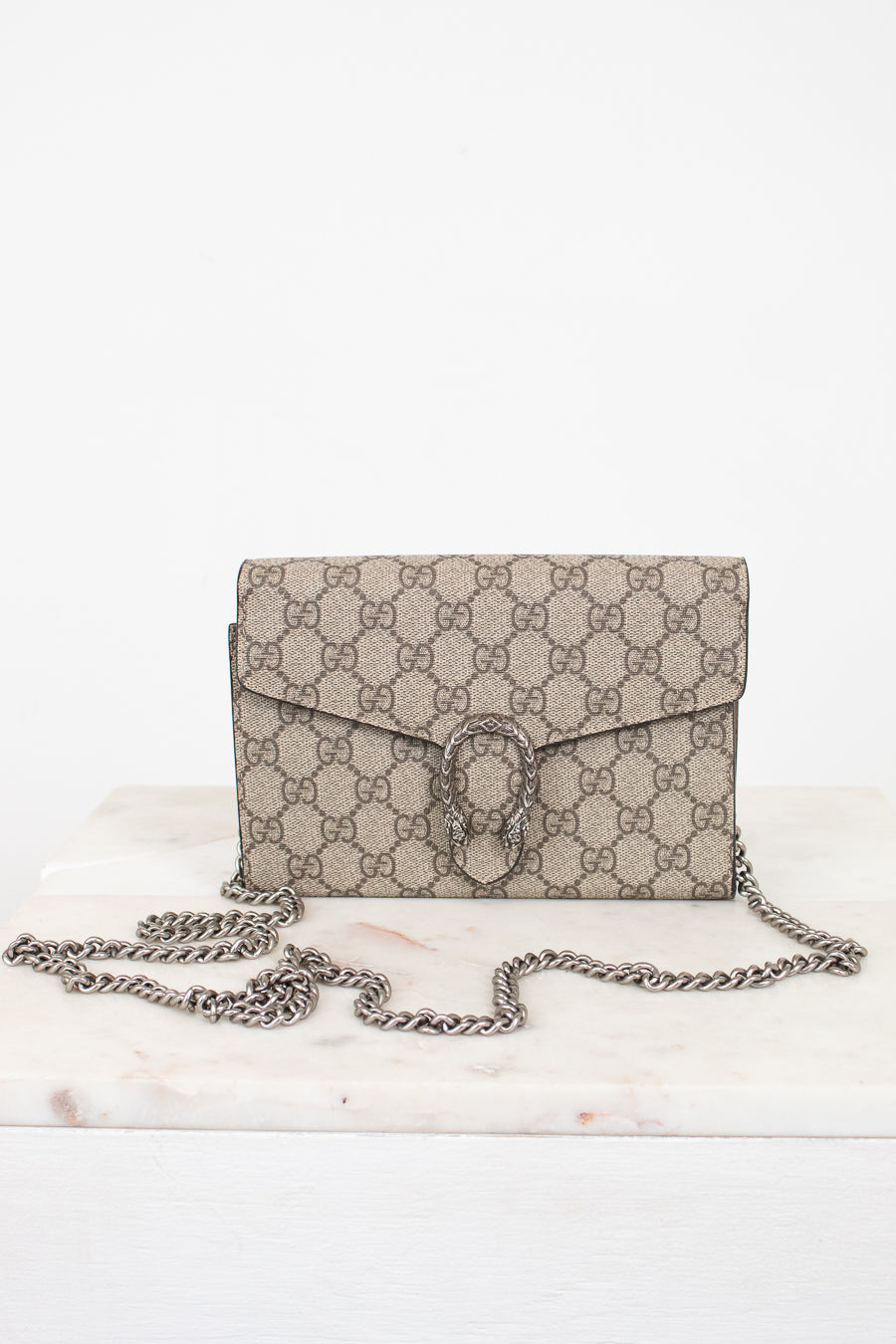 A beige and brown GG Supreme coated canvas shoulder bag with a silver-tone chain strap, displayed on a white marble surface against a white background.