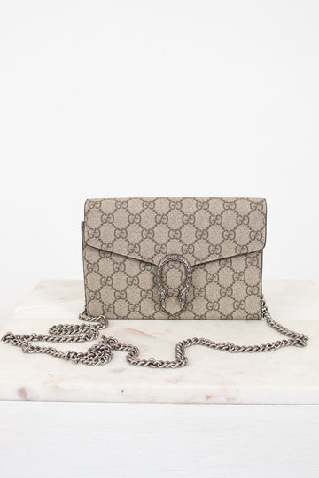 A beige and brown GG Supreme coated canvas shoulder bag with a silver-tone chain strap, displayed on a white marble surface against a white background.