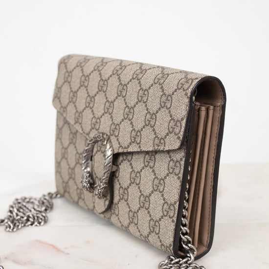 A Gucci shoulder bag in beige and brown GG Supreme canvas with a silver-tone clasp and chain strap, resting on a light marble surface against a white background.