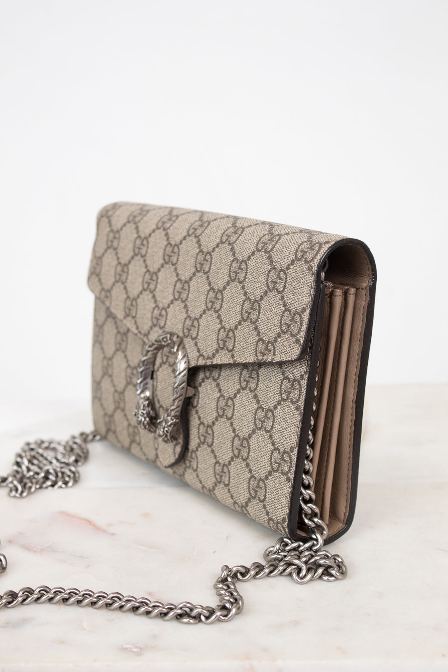 A Gucci shoulder bag in beige and brown GG Supreme canvas with a silver-tone clasp and chain strap, resting on a light marble surface against a white background.