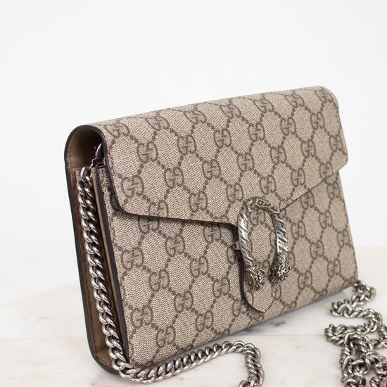 A Gucci shoulder bag in beige GG Supreme canvas with brown leather trim and a silver-tone chain strap, displayed on a light marble surface against a white background.