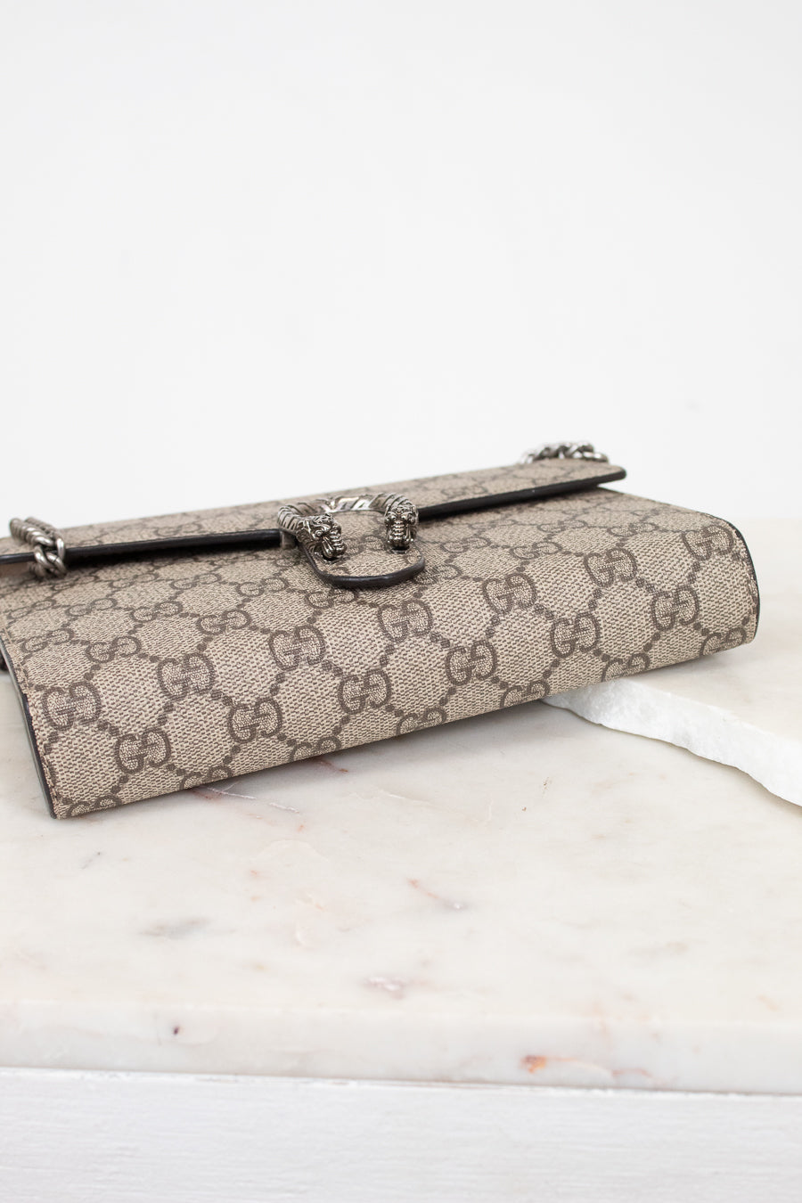 A Gucci monogram canvas clutch bag with a silver-tone horsebit clasp, resting on a white marble surface against a white background.