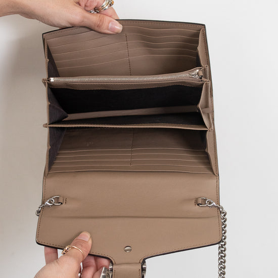 A taupe-brown leather interior of handbag shown open, held by two hands to display its interior compartments, including card slots, a zippered pocket, and a main compartment. The bag features a silver-tone chain strap and is photographed against a clean, off-white background.