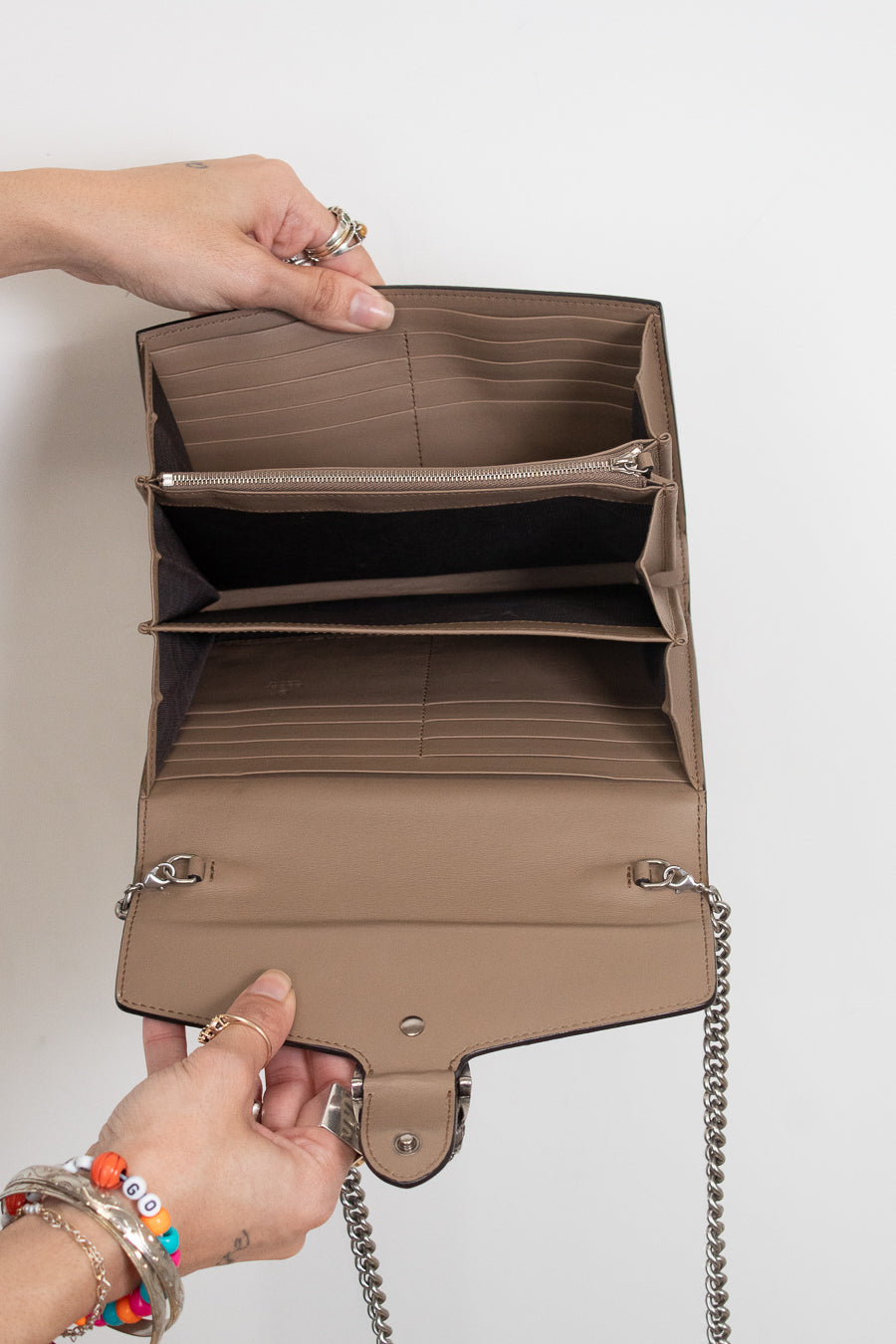 A taupe-brown leather interior of handbag shown open, held by two hands to display its interior compartments, including card slots, a zippered pocket, and a main compartment. The bag features a silver-tone chain strap and is photographed against a clean, off-white background.