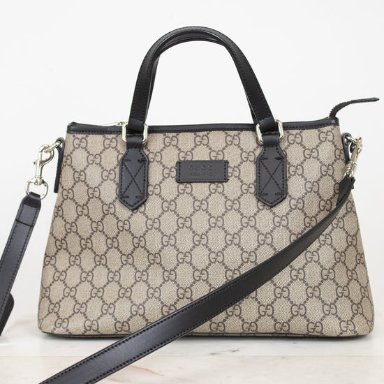 A beige and brown GG Supreme monogram handbag with black leather handles, trim, and a detachable shoulder strap, displayed on a light marble surface against a white background.