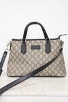 A beige and brown GG Supreme monogram handbag with black leather handles, trim, and a detachable shoulder strap, displayed on a light marble surface against a white background.