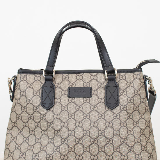 A beige and brown monogram handbag with black leather handles and trim, featuring the interlocking GG pattern and a central logo plaque.