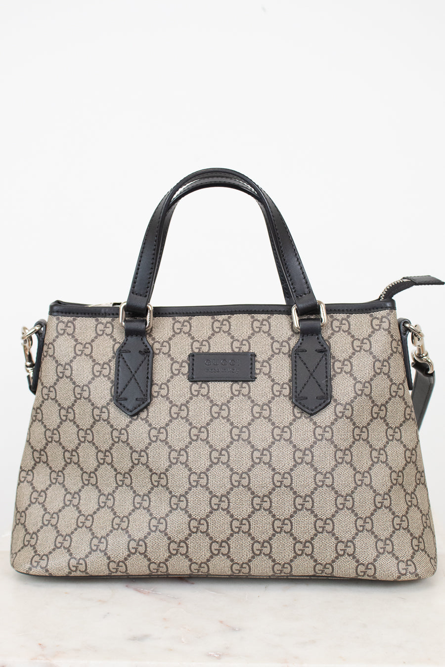 A beige and brown monogram handbag with black leather handles and trim, featuring the interlocking GG pattern and a central logo plaque.