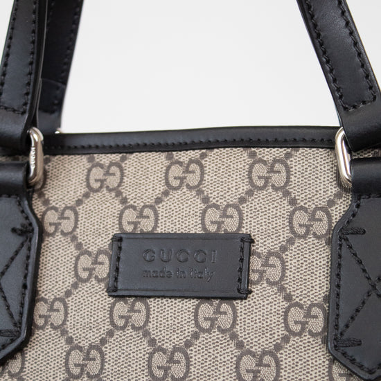 Close-up of a Gucci handbag featuring the brand's signature GG monogram canvas in beige and brown, accented by black leather trim and handles with silver-tone hardware.