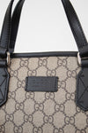 Close-up of a Gucci handbag featuring the brand's signature GG monogram canvas in beige and brown, accented by black leather trim and handles with silver-tone hardware.