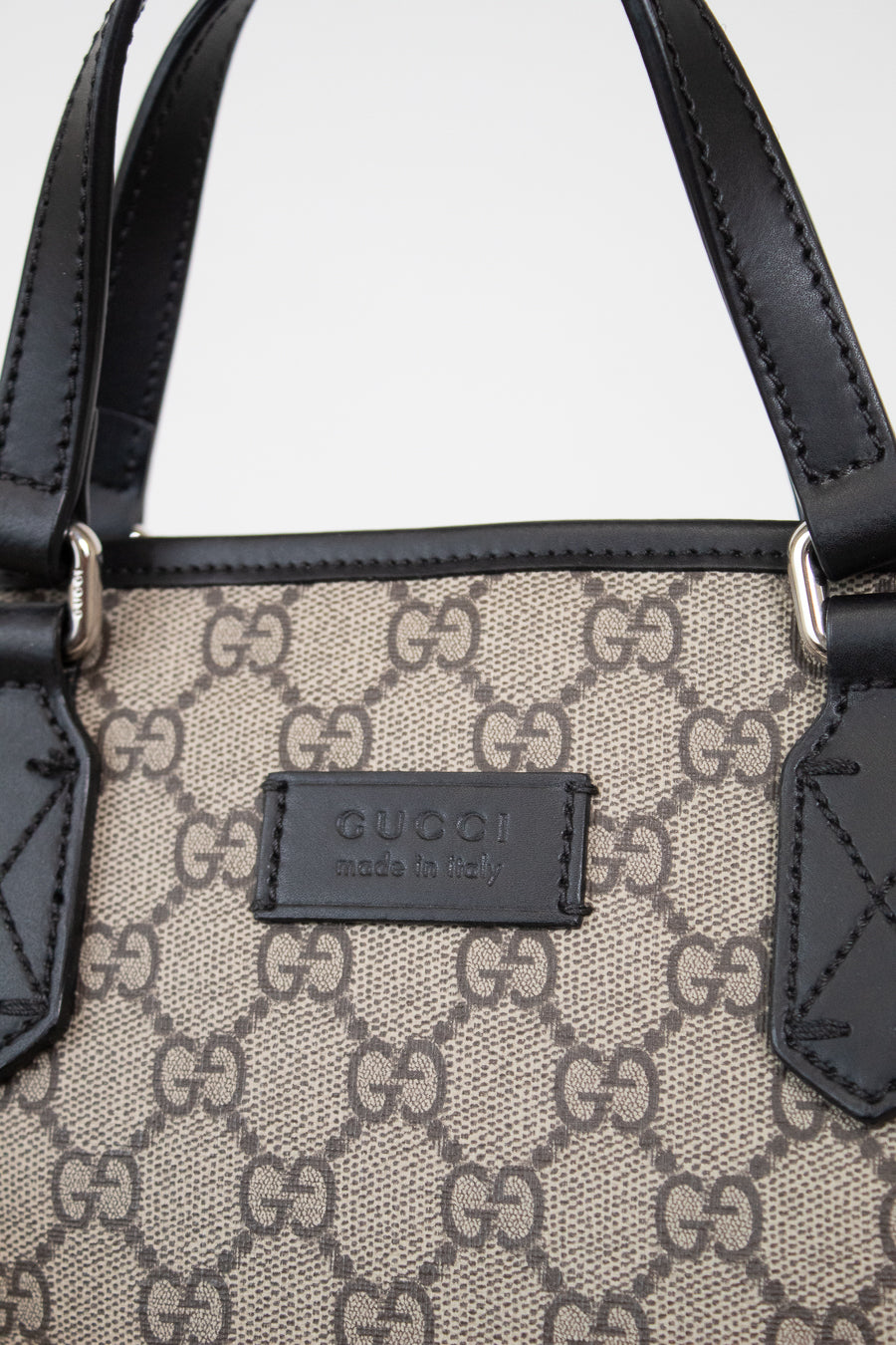 Close-up of a Gucci handbag featuring the brand's signature GG monogram canvas in beige and brown, accented by black leather trim and handles with silver-tone hardware.