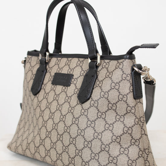 A beige and brown GG Supreme canvas handbag with black leather handles and trim, shown on a light marble surface against a white background.