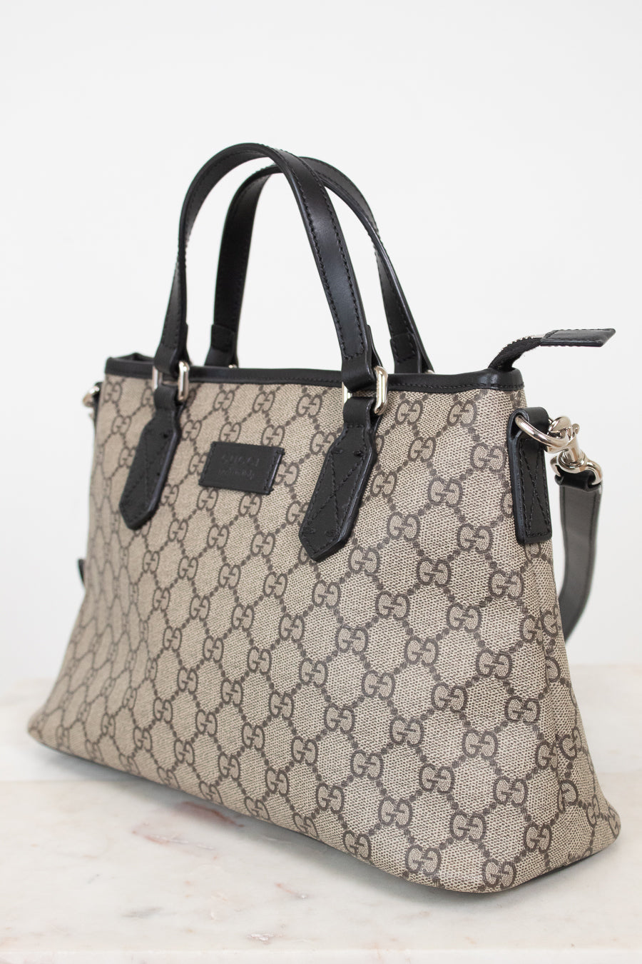 A beige and brown GG Supreme canvas handbag with black leather handles and trim, shown on a light marble surface against a white background.