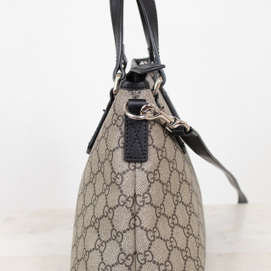 A small Gucci handbag in beige and brown GG Supreme canvas with black leather handles and trim, shown from the side on a light marble surface against a white background.