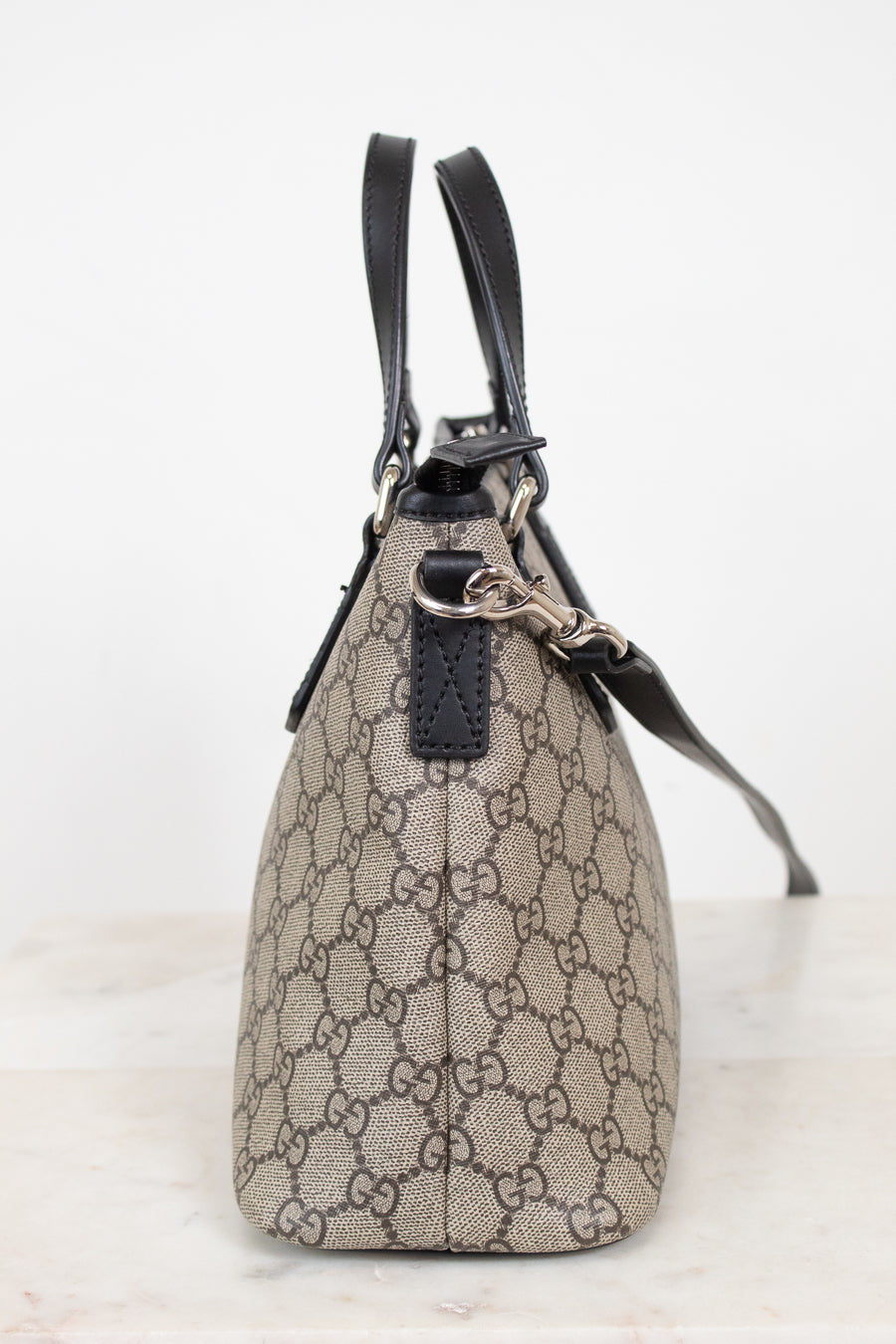 A small Gucci handbag in beige and brown GG Supreme canvas with black leather handles and trim, shown from the side on a light marble surface against a white background.