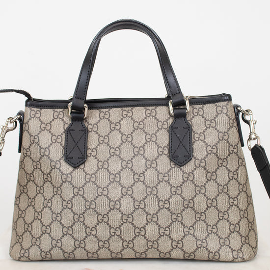 A Gucci handbag in beige and brown GG Supreme canvas with black leather handles and trim, displayed on a light marble surface against a white background.